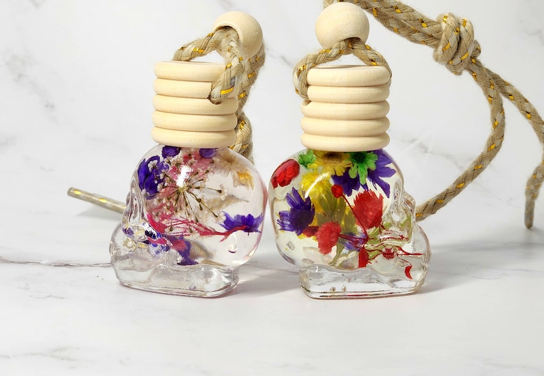 May include: Two clear, skull-shaped car air fresheners filled with colourful dried flowers. Each has a wooden cap and a braided rope for hanging. The flowers include purple, red, and yellow blooms.