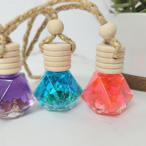 May include: Three small glass bottles with wooden stoppers and a gold string hanging from each. The bottles are filled with colorful liquids and glitter. The bottles are purple, blue, and pink.