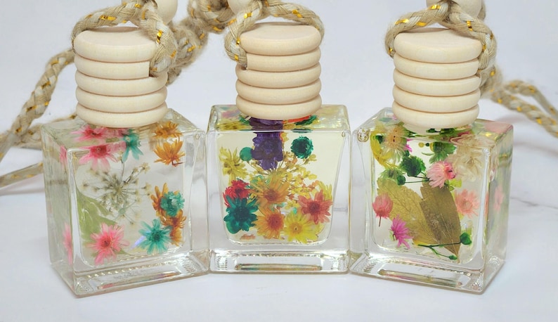 May include: Three clear, square glass bottles filled with colourful dried flowers. Each bottle has a light wood cap and a braided rope for hanging. The flowers include pink, yellow, and blue blooms.