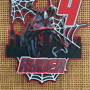 Puede incluir: Adorno para tarta en capas con un superh&eacute;roe con un traje negro y rojo, con el n&uacute;mero 4 rojo. El nombre "RYDER" se muestra en letras plateadas con purpurina. El dise&ntilde;o incluye telara&ntilde;as y una silueta de ciudad.