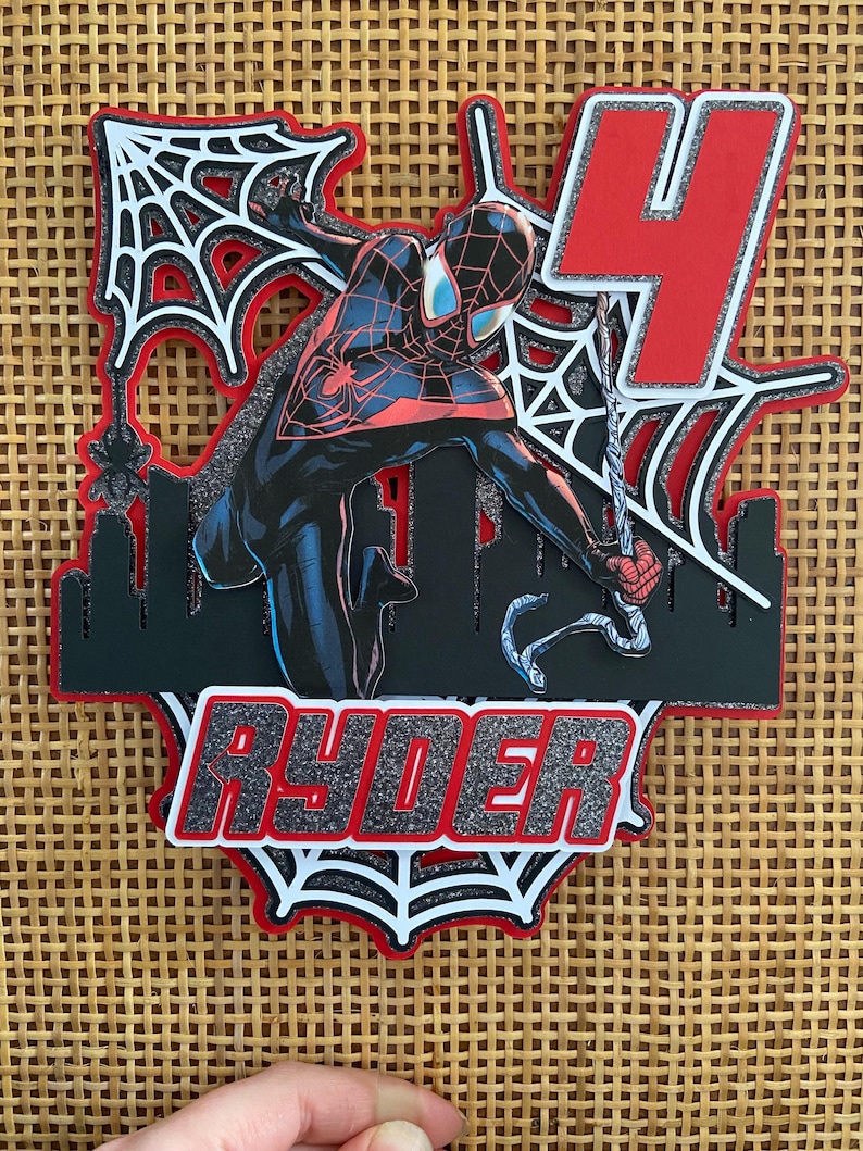 Puede incluir: Adorno para tarta en capas con un superh&eacute;roe con un traje negro y rojo, con el n&uacute;mero 4 rojo. El nombre "RYDER" se muestra en letras plateadas con purpurina. El dise&ntilde;o incluye telara&ntilde;as y una silueta de ciudad.