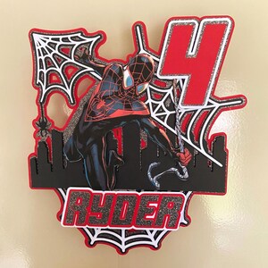 Puede incluir: Un adorno para tarta en capas con un personaje de Spider-Man en una pose din&aacute;mica, con una combinaci&oacute;n de colores rojo y negro. El n&uacute;mero "4" se muestra de forma destacada, junto con el nombre "Ryder". El dise&ntilde;o incluye telara&ntilde;as y una silueta de ciudad.