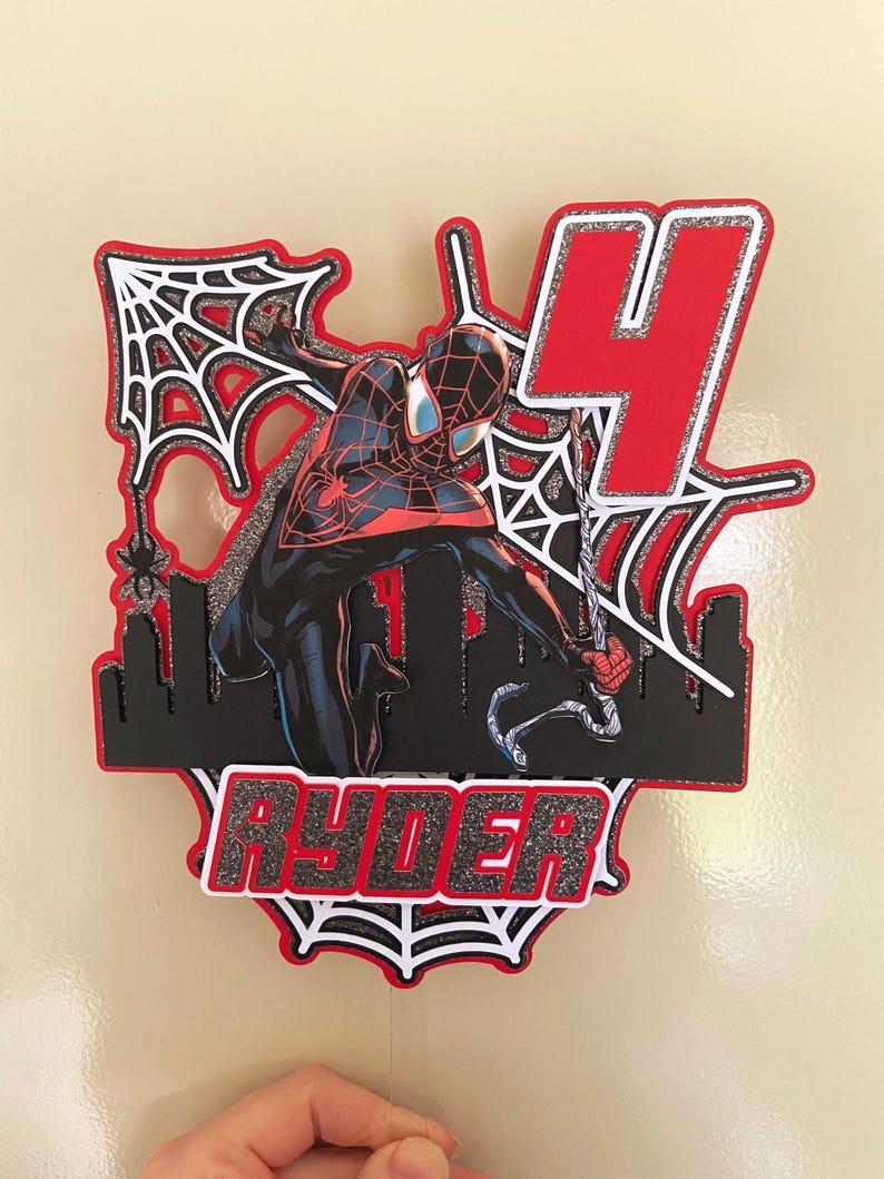 Puede incluir: Un adorno para tarta en capas con un personaje de Spider-Man en una pose din&aacute;mica, con una combinaci&oacute;n de colores rojo y negro. El n&uacute;mero "4" se muestra de forma destacada, junto con el nombre "Ryder". El dise&ntilde;o incluye telara&ntilde;as y una silueta de ciudad.