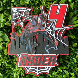 Puede incluir: Un adorno para pastel en capas con un personaje de Spider-Man en una pose din&aacute;mica, con una combinaci&oacute;n de colores rojo y negro. El dise&ntilde;o incluye una silueta de ciudad, un gran n&uacute;mero 4 y el nombre "RYDER" en letras en negrita, todo sobre un fondo de follaje verde.