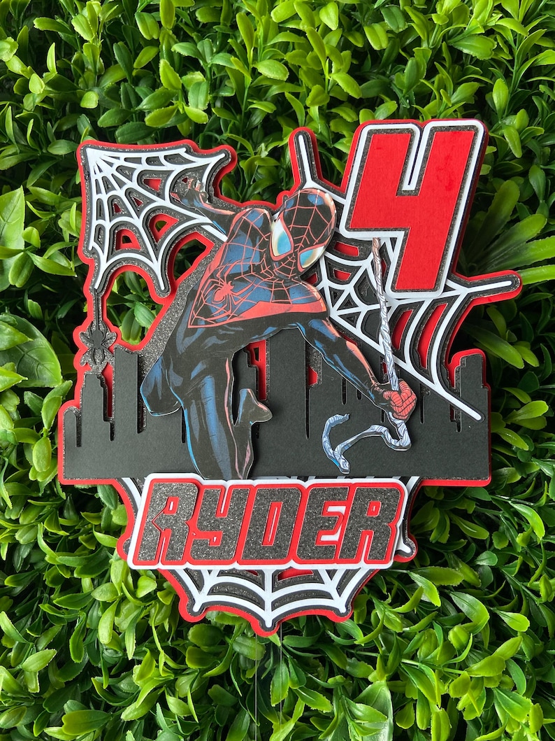Puede incluir: Un adorno para pastel en capas con un personaje de Spider-Man en una pose din&aacute;mica, con una combinaci&oacute;n de colores rojo y negro. El dise&ntilde;o incluye una silueta de ciudad, un gran n&uacute;mero 4 y el nombre "RYDER" en letras en negrita, todo sobre un fondo de follaje verde.
