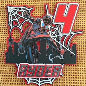 Puede incluir: Adorno para tarta en capas con Spider-Man en una pose din&aacute;mica, con un n&uacute;mero 4 rojo y el nombre "RYDER". El dise&ntilde;o incluye una silueta de ciudad y telara&ntilde;as, todo delineado en rojo y negro.