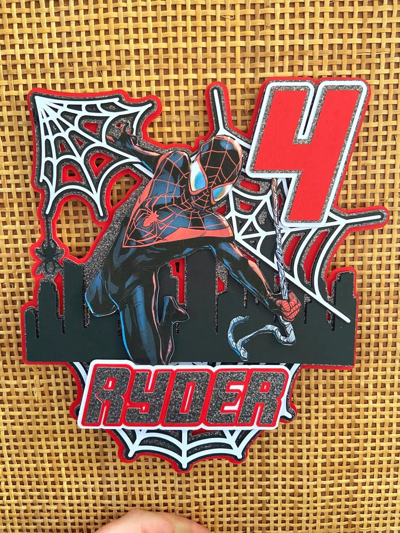 Puede incluir: Adorno para tarta en capas con Spider-Man en una pose din&aacute;mica, con un n&uacute;mero 4 rojo y el nombre "RYDER". El dise&ntilde;o incluye una silueta de ciudad y telara&ntilde;as, todo delineado en rojo y negro.