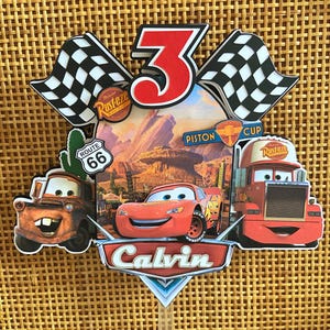 Op de afbeelding: Een kleurrijke Cars-thema taarttopper met Bliksem McQueen, Takel en Mack. Het ontwerp bevat een rood nummer 3, geblokte vlaggen en de naam "Calvin". De achtergrond toont een woestijnlandschap.