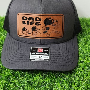 May include: A gray trucker hat with a black mesh back and a brown leather patch. The patch features the words "DAD LIFE" above a cartoon dog family. The hat has an adjustable strap and a curved brim.