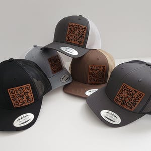 May include: A collection of baseball caps in various colors, including black, gray, and brown. Each cap features a QR code patch and a mesh back. The caps are displayed on a white surface.