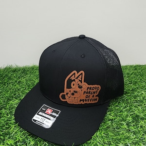 May include: Black trucker hat with a brown patch featuring a cartoon dog and the text "Proud Parent of a Muffin". The hat is made by Richardson 112.