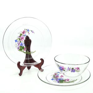 May include: A clear glass dish set featuring a plate, a bowl, and a smaller plate. Each piece is decorated with a floral design in shades of purple, pink, and blue. The plate is displayed on a dark brown wooden stand.