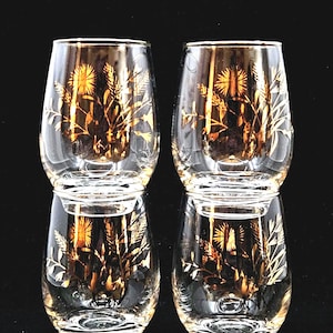 Könnte beinhalten: Vier klare, stielose Gläser mit einem goldenen Blumendesign. Die Gläser sind paarweise gestapelt, wobei die unteren beiden die oberen beiden widerspiegeln. Das goldene Design zeigt Weizen und andere florale Elemente.
