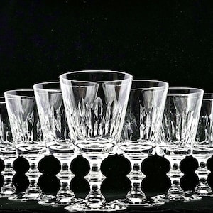 May include: A collection of eight clear crystal wine glasses with intricate cut patterns. The glasses have a classic design with a stemmed base and a flared bowl. The glasses are arranged in a row against a black background.