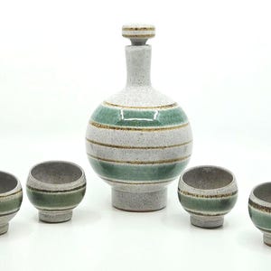 May include: A ceramic decanter set with a round body and a stopper, accompanied by four small cups. The set features a speckled gray base with horizontal stripes of green and brown. The cups have a similar design.