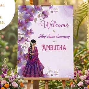 May include: A digital download wedding sign for a Half Saree Ceremony. The sign features a floral design with the name "AMRUTHA". The sign is displayed on a wooden easel, surrounded by floral arrangements and greenery.