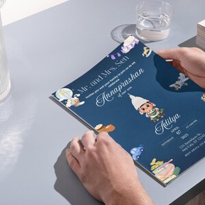 May include: A dark blue rectangular invitation card featuring a cartoon illustration of a child in traditional attire, surrounded by small icons of toys and a hot air balloon. The text reads Mr. and Mrs. Sen invite you to celebrate Annaprashan of our son Aditya, Saturday April 07 2025, 123 Anywhere St, Any City, ST 12345.