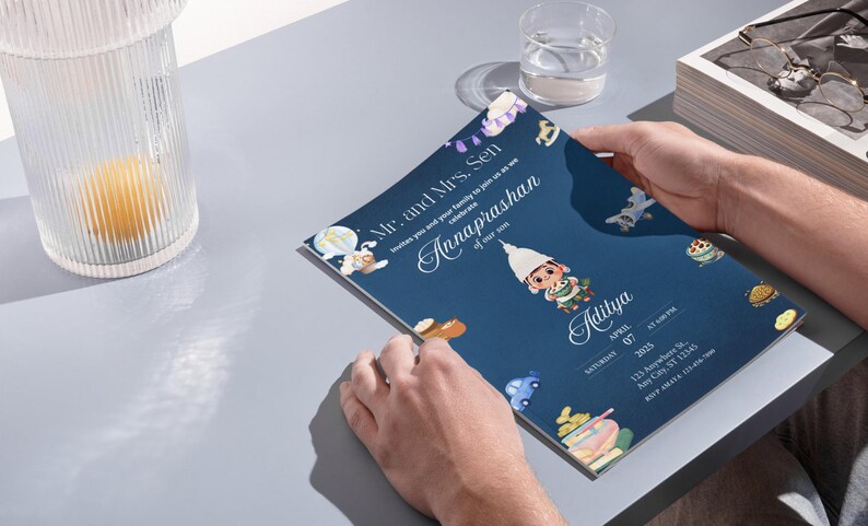 May include: A dark blue rectangular invitation card featuring a cartoon illustration of a child in traditional attire, surrounded by small icons of toys and a hot air balloon. The text reads Mr. and Mrs. Sen invite you to celebrate Annaprashan of our son Aditya, Saturday April 07 2025, 123 Anywhere St, Any City, ST 12345.