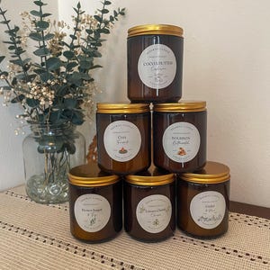 May include: A collection of six brown glass jar candles with gold lids. Each candle has a white label with black text, including scents like "Cocoa Butter" and "Bourbon Butterscotch". The candles are arranged on a table with a neutral-toned tablecloth.