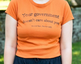 May include: Orange t-shirt with the text "Your government doesn't care about you. Never has. Never will." The shirt has a crew neck and short sleeves. The person is wearing dark blue cargo pants.