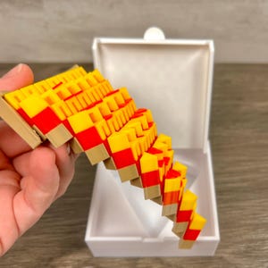 May include: A colorful, geometric sculpture with a cascade design. The sculpture features a repeating pattern of yellow, red, and tan blocks. It is held in front of a white box, suggesting it may be a piece of jewelry or a decorative item.