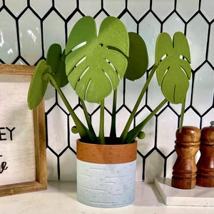 May include: A decorative artificial monstera plant in a two-tone ceramic pot. The pot has a light blue textured base and a brown rim. The plant features large, green, stylized leaves and green stems. The background includes a white and black tile pattern.