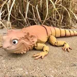 May include: A beige and brown articulated toy lizard with a large head and a friendly expression. The lizard has a segmented tail and is posed on a light brown surface.