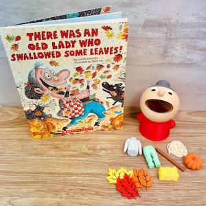 May include: A children's book titled There Was An Old Lady Who Swallowed Some Leaves by Lucille Colandro, alongside a beige figurine with a wide open mouth, a grey shirt, green pants, a brown stick, a white rope, a pumpkin, and leaf cutouts.