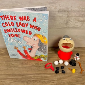 May include: A children's book titled There Was a Cold Lady Who Swallowed Some Snow, featuring a character with a red shirt and yellow hat. Beside it is a red and beige plastic figure with an open mouth, accompanied by small accessories including white spheres, black pieces, a black hat, a red ring, a yellow tool, and a small snowman figure.