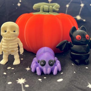 May include: A collection of five Halloween-themed figurines: a pumpkin, a skeleton, a bat, a mummy, and a spider. The figurines are made of plastic and are painted in bright colors. The pumpkin is orange, the skeleton is white, the bat is black, the mummy is beige, and the spider is purple.