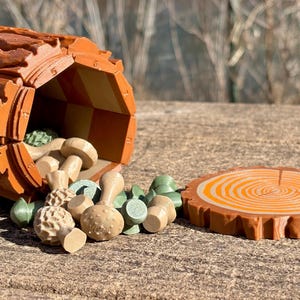 Op de afbeelding: Een houten speelgoedset met een holle boomstam met een dambordinterieur, waaruit verschillende paddenstoelvormige stukken vallen. Een apart, rond houten stuk lijkt op een schijf van een boom. De set is in tinten bruin, beige en groen.