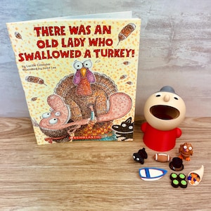 May include: A children's book titled There Was An Old Lady Who Swallowed A Turkey by Lucille Colandro, featuring a turkey illustration. Beside it sits a beige and red plastic character figure with a wide open mouth and small toy accessories.