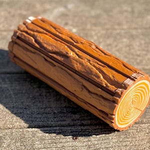 Op de afbeelding: Een decoratieve log-vormige doos met een gedetailleerde houtnerftextuur. De uiteinden van de log hebben concentrische cirkels en de algehele kleur is een warm bruin met lichtere accenten. De doos is ontworpen om op een realistische log te lijken.