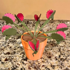 May include: A plastic toy plant with green stems and pink flower-like traps. The plant is in a terracotta pot.