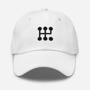May include: White baseball cap with a black graphic of a gear shift pattern on the front. The cap has a curved brim and an adjustable strap at the back. The design is simple and modern.
