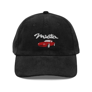 Puede incluir: Gorra de béisbol de pana negra con la palabra "Miata" bordada en escritura blanca sobre un gráfico de coche rojo. La gorra tiene una visera curva y un botón en la parte superior.