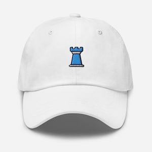 Puede incluir: Gorra de béisbol blanca con una pieza de ajedrez de torre bordada en azul y blanco. La gorra tiene una visera curva y una correa ajustable. El diseño de la torre es un símbolo clásico del ajedrez.