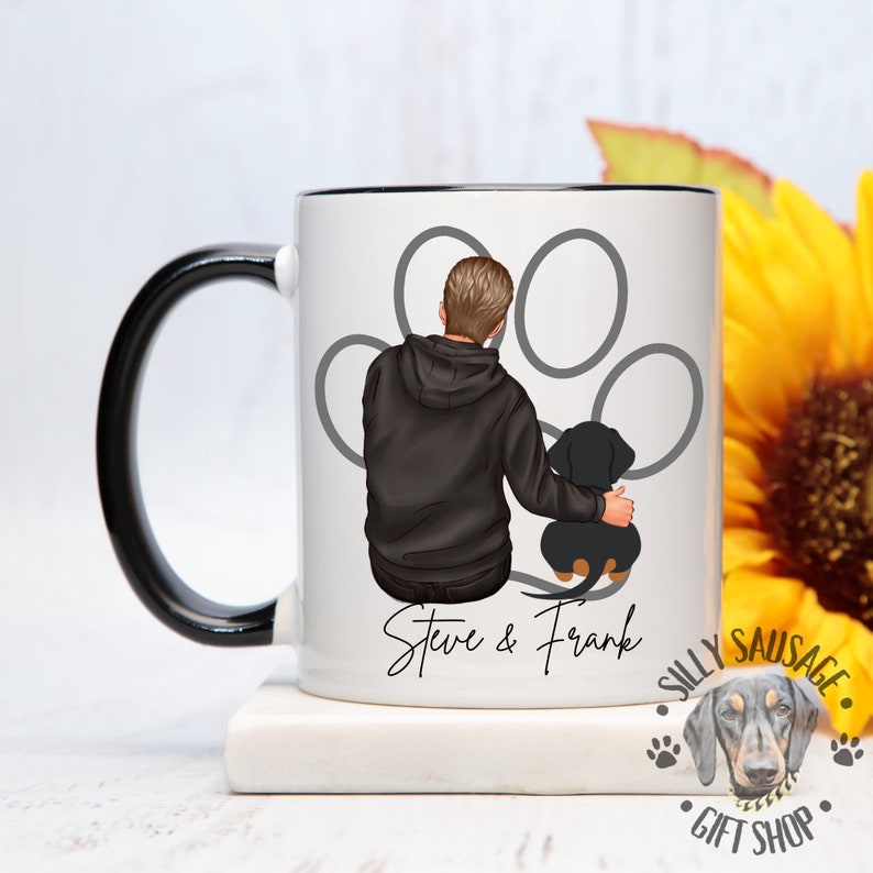 Puede incluir: Taza de cer&aacute;mica blanca con asa y borde negros, con una ilustraci&oacute;n de una persona con un perro salchicha dentro de una huella de pata. La taza tiene el texto "Steve & Frank" y un logotipo que dice "Silly Sausage Gift Shop".