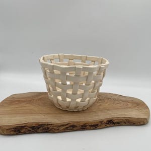 以下が含まれることがあります： 円筒形の小さなクリーム色の織りバスケット。バスケットは、自然なエッジを持つ木製の楕円形のまな板の上に置かれています。背景は無地の白です。