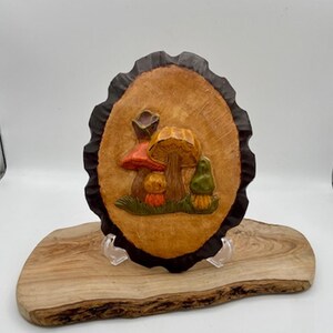 May include: A wooden, oval-shaped wall hanging featuring a relief carving of several mushrooms in shades of orange, red, and green. The mushrooms are set against a light brown background with a dark brown, scalloped edge. The piece is displayed on a wooden base.