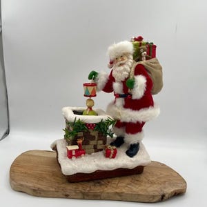 May include: A Christmas-themed figurine of Santa Claus in a red suit with white faux fur trim, holding a drum and a sack of gifts. The figurine stands on a snowy base with a chimney and a toy train. The base is set on a wooden board.
