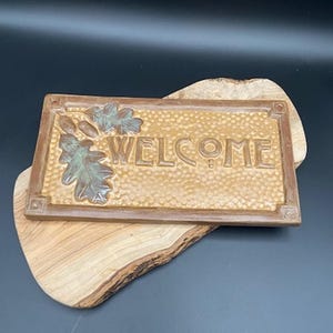 May include: A rectangular brown welcome sign with the word "WELCOME" in raised letters. The sign features an oak leaf design and is set on a wooden base. The sign is made of a textured material.
