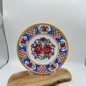 Op de afbeelding: Een wit keramiek bord met een geel, blauw en paars bloemenontwerp. Het bord heeft een gele rand en een geruit patroon rond de rand. Het bord staat op een houten standaard.