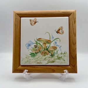 Puede incluir: Un azulejo de cerámica enmarcado con una pintura de acuarela de setas y mariposas. El azulejo está rodeado de un marco de madera y se encuentra sobre una superficie blanca.