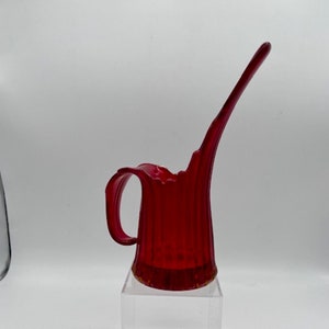 May include: A red glass pitcher with a unique, curved spout and a handle. The pitcher has a ribbed texture and is sitting on a clear acrylic stand.
