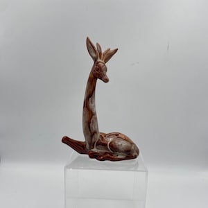 May include: A brown and white ceramic deer figurine in a resting pose. The deer has a long neck and detailed antlers. The figurine is displayed on a clear acrylic stand against a white background.