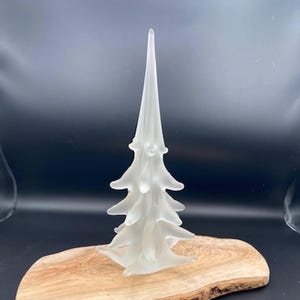 Könnte beinhalten: Eine Weihnachtsbaumskulptur aus mattiertem Glas mit spitzer Spitze und geschichteten Ästen. Der Baum wird auf einem Schneidebrett aus Naturholz mit dunklem Hintergrund präsentiert. Der Baum ist etwa 20 cm hoch.