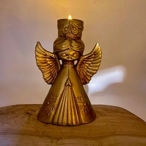 May include: A gold-colored angel candle holder with a lit candle. The angel has detailed wings and a crown. The candle holder is placed on a wooden surface, creating a warm and inviting ambiance. The angel's design is vintage-inspired.