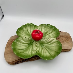 Puede incluir: Un plato de cerámica verde con forma de trébol de cuatro hojas, con una pieza central roja en forma de tomate. El plato está colocado sobre una tabla de cortar de madera, creando una presentación rústica y elegante.