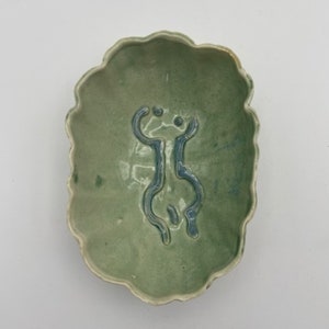 May include: A green ceramic dish with a wavy edge and a stylised human figure design in blue glaze.